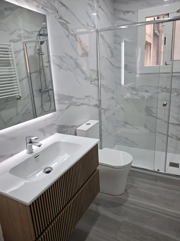 Baño completo con revestimiento efecto mármol, lavabo con mueble de madera y ducha acristalada — Junior Reformas